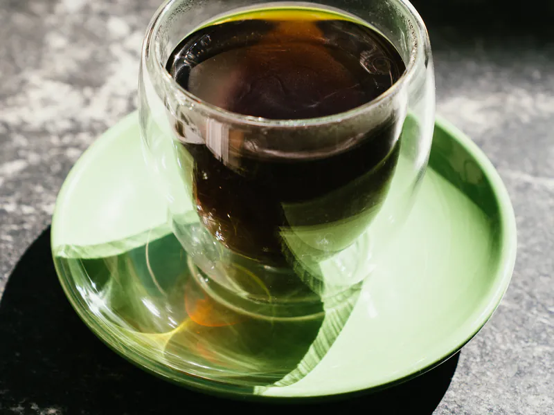 Café de especialidad negro en vaso de vidrio sobre plato verde, con luz de la tarde